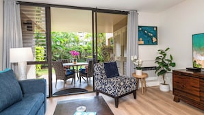 A living room with a sofa, a coffee table, a patterned armchair, a small side table, a potted plant, and a sliding glass door leading to an outdoor area with a table and chairs. - Charming Beachside Garden Vistas (Kihei)