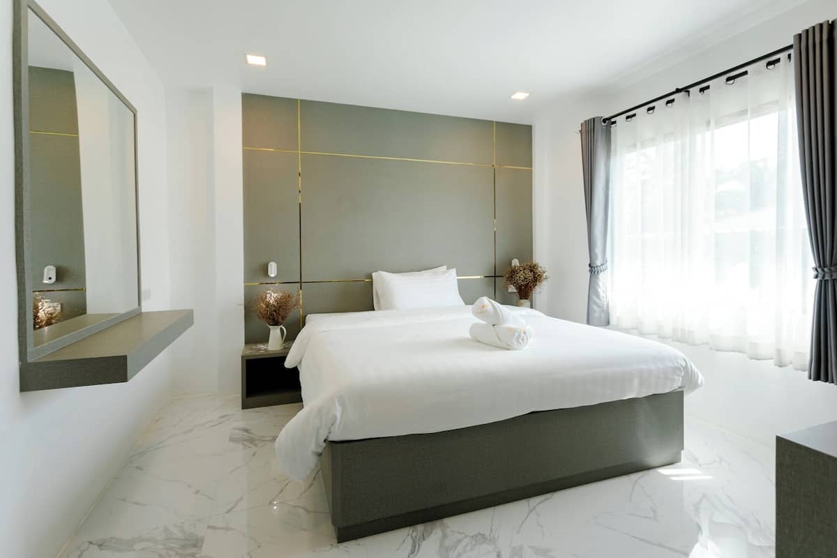 A modern hotel room with a large bed, a bedside table with a vase of dried flowers, a mirror, and a view of the city through large windows.