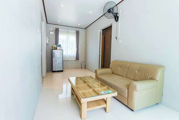 A modern living room with a beige sofa, a glass-top coffee table, and a compact kitchenette in the background. - The Neenlawat Riverside Resort & Spa (Phunphin)