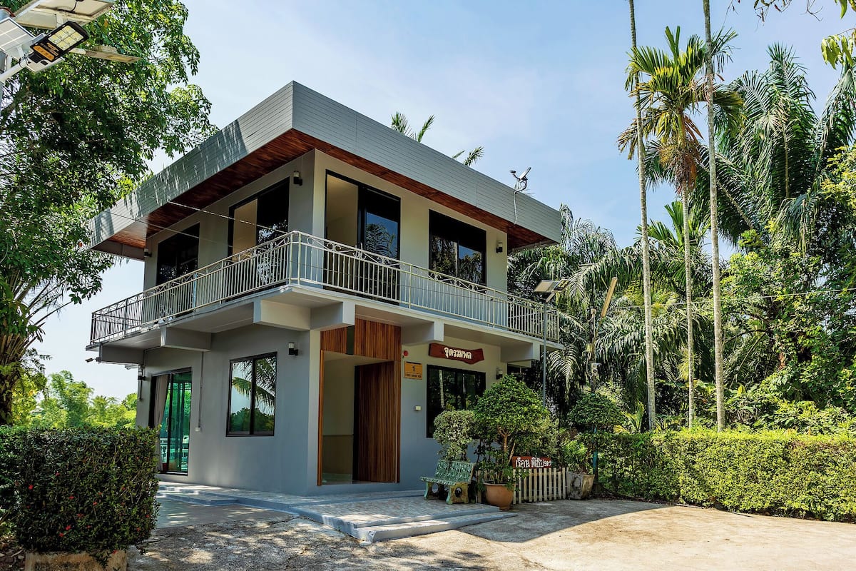 A modern two-story building with a balcony, surrounded by lush greenery and palm trees.