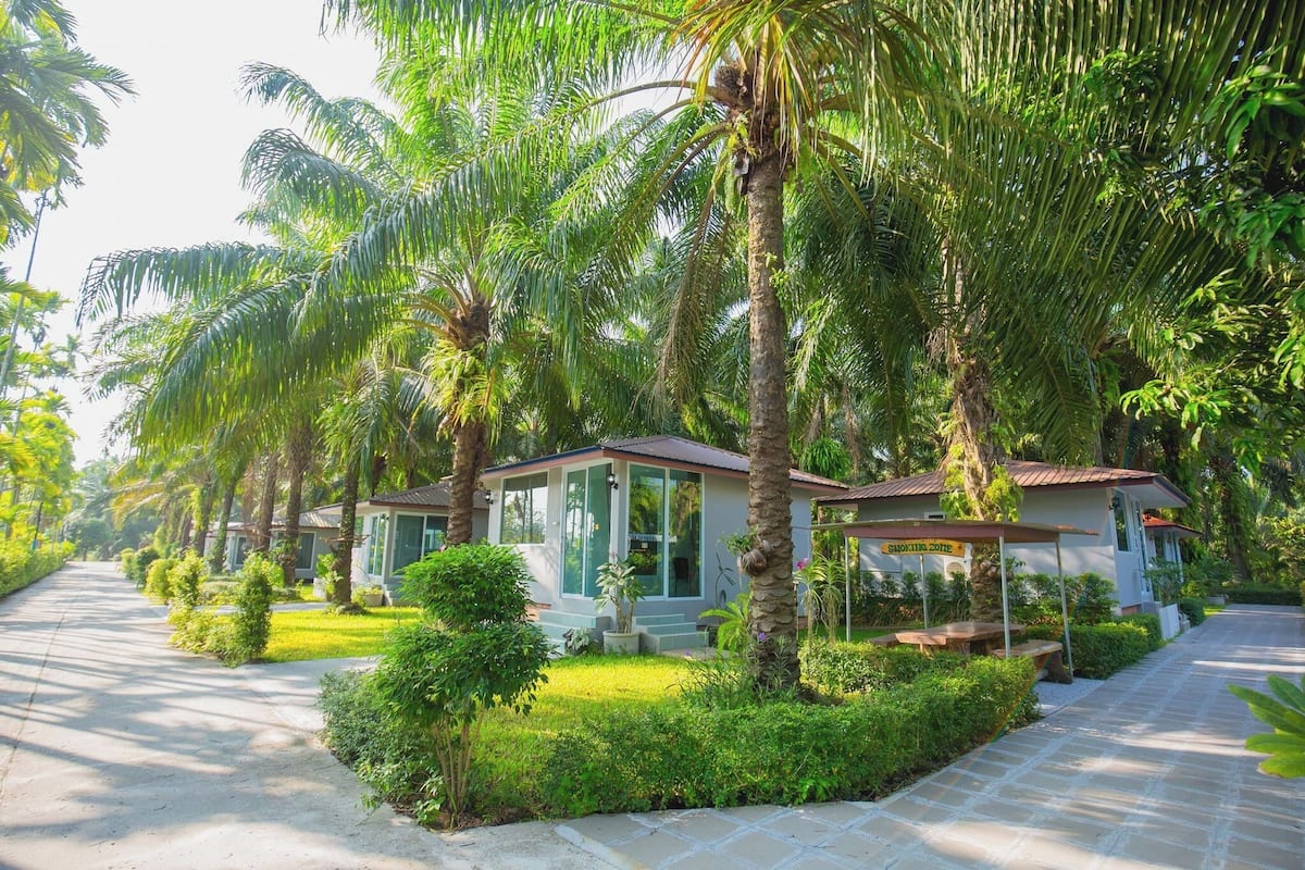 A row of two-story villas with a paved driveway, surrounded by lush greenery and palm trees.