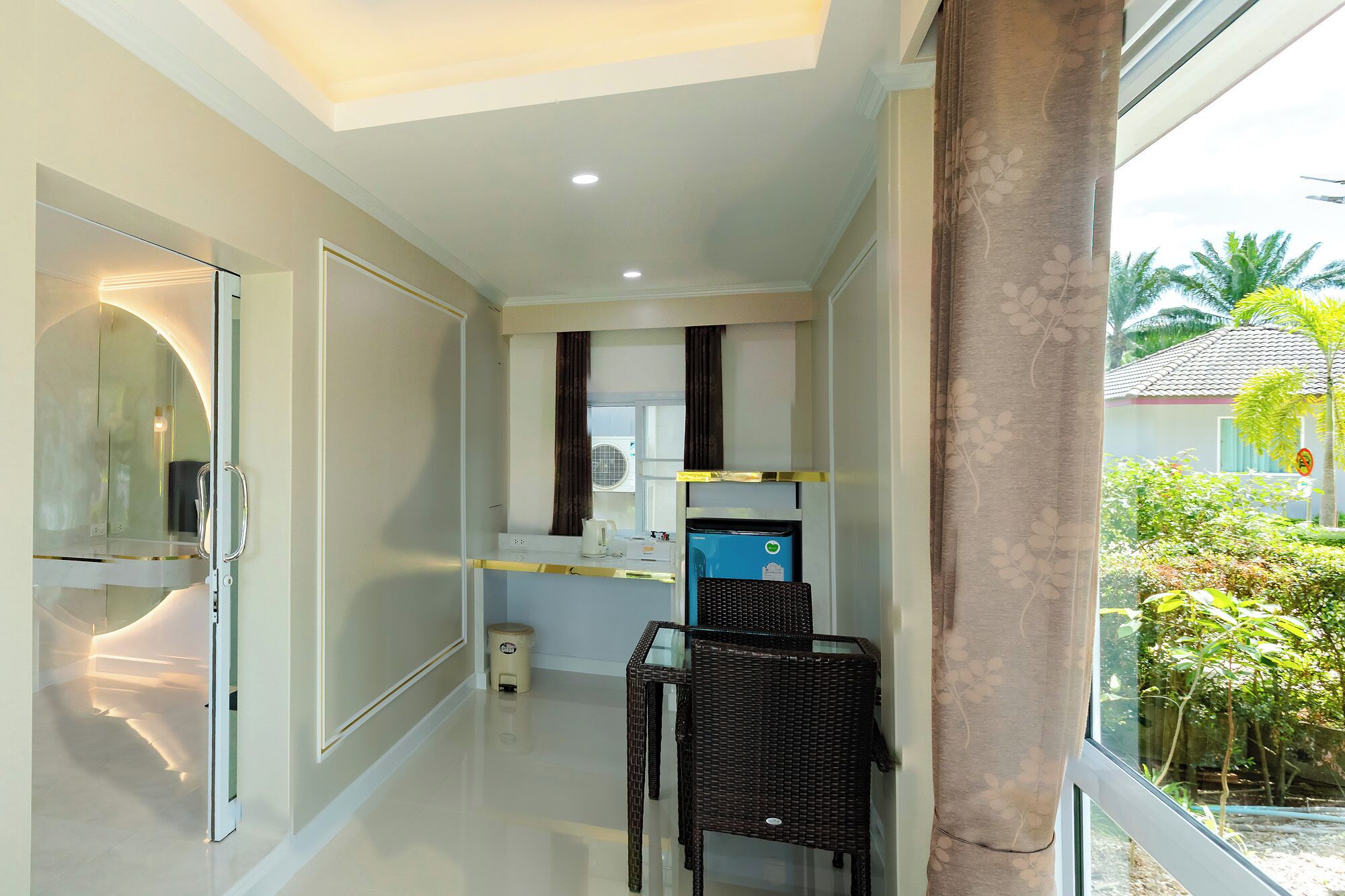 A modern hotel room with a glass door leading to a balcony, a kitchenette with a coffee machine, and a wicker chair.