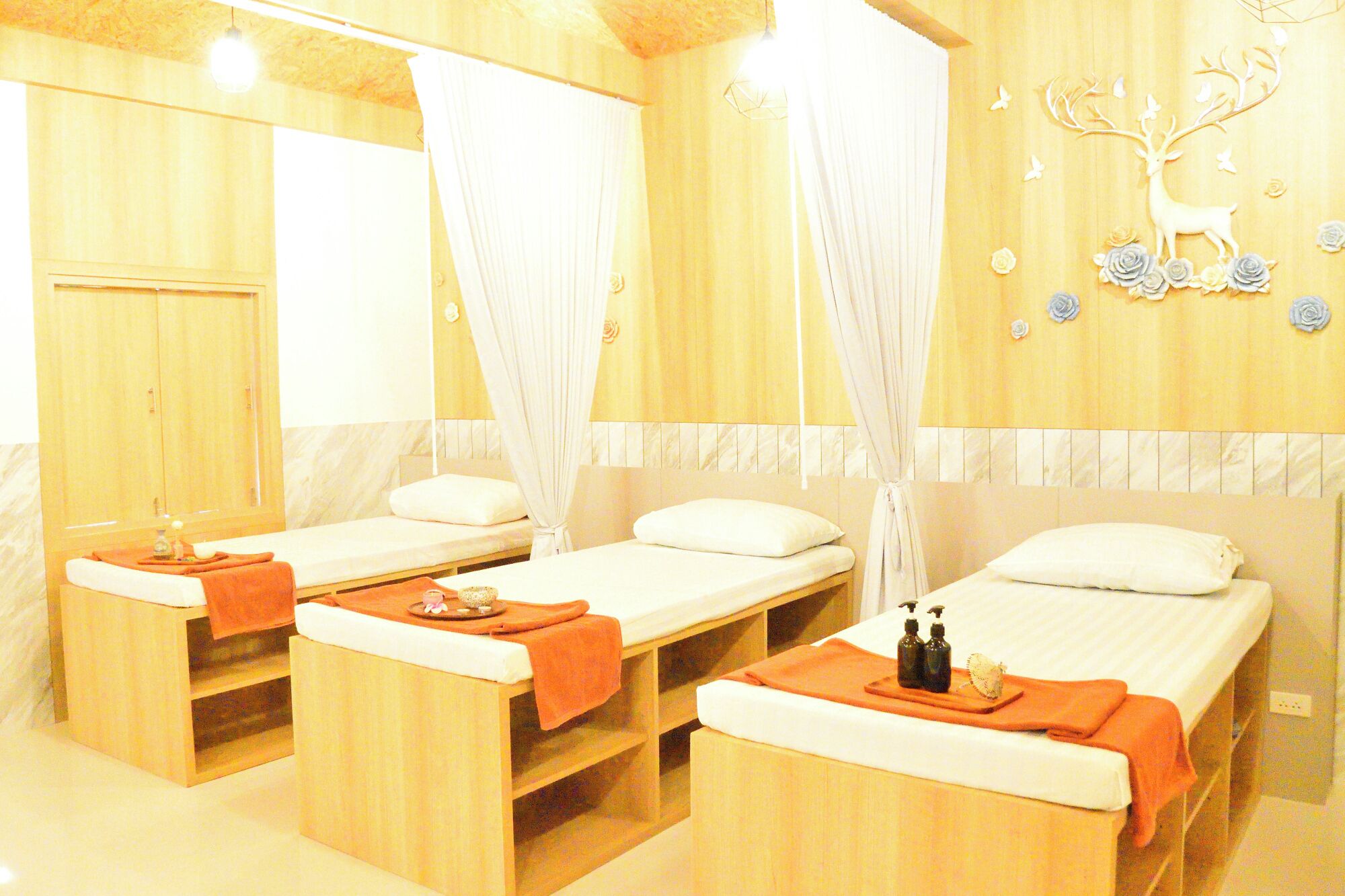 A room with three wooden beds, each with a white pillow and an orange towel. There is a wooden cabinet with a mirror and a wooden shelf with a bottle.