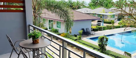 A balcony with a table and chairs, overlooking a swimming pool and a building with a tiled roof.