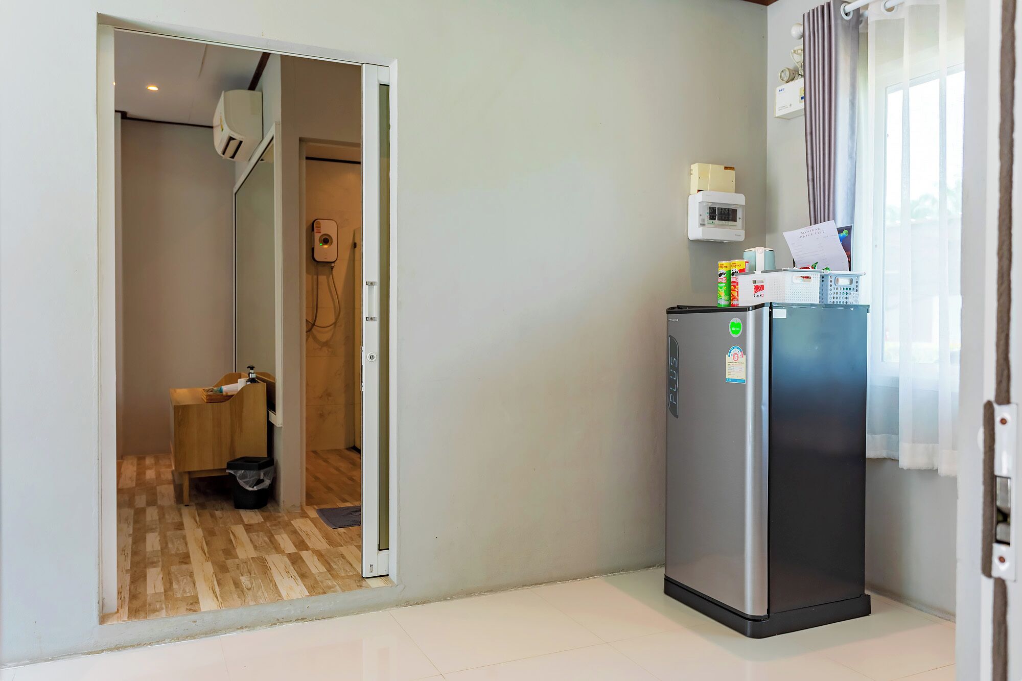 A compact kitchen with a refrigerator, a microwave, and a small countertop. A wooden cabinet and a trash bin are visible in the adjacent room.