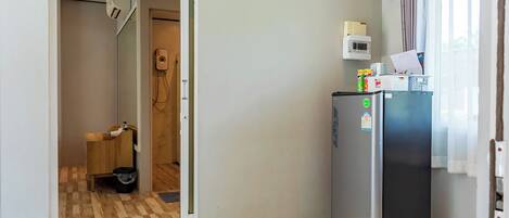 A compact kitchen with a refrigerator, a microwave, and a small countertop. A wooden cabinet and a trash bin are visible in the adjacent room.