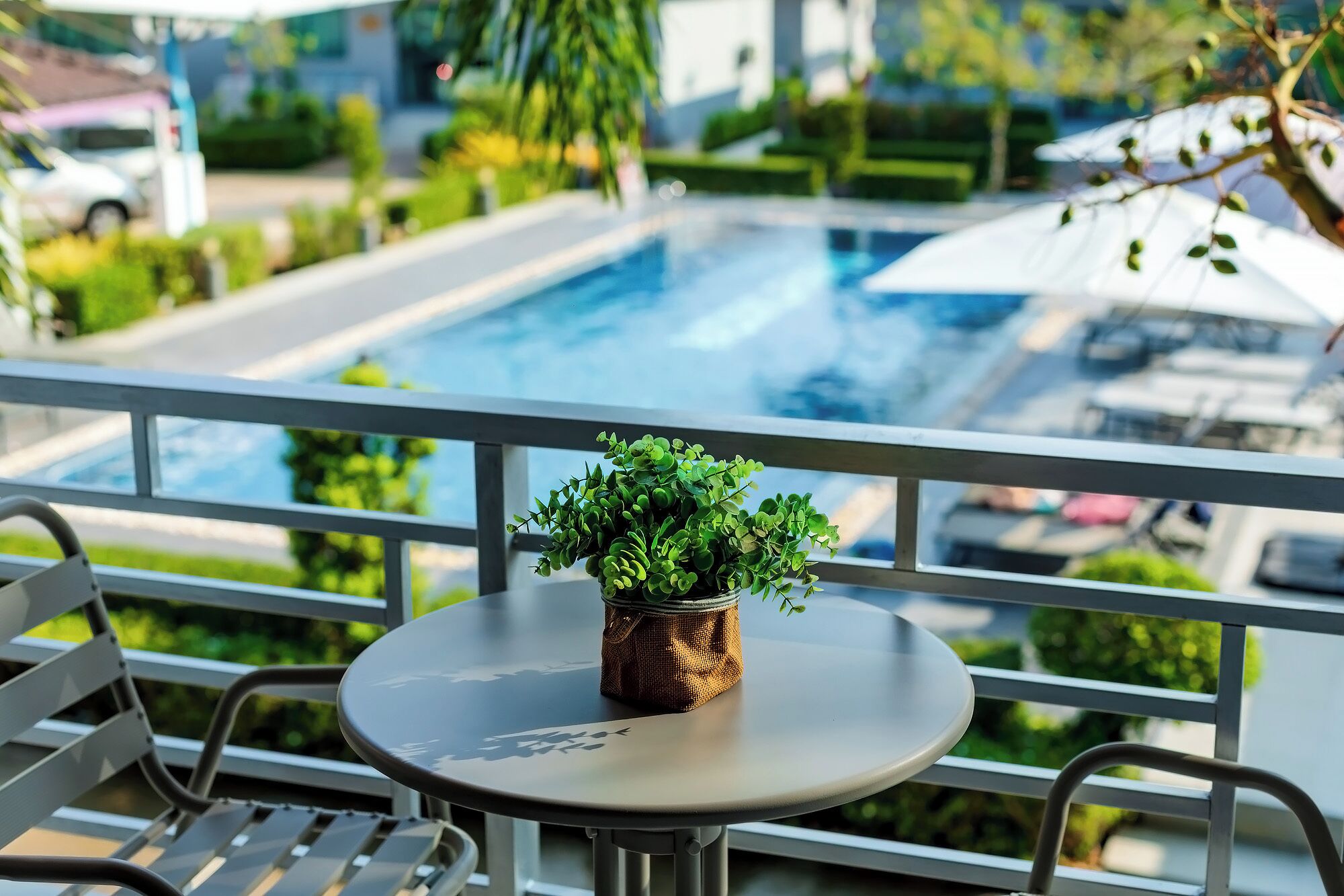 A balcony with a small table and a potted plant, overlooking a swimming pool and a parking area.