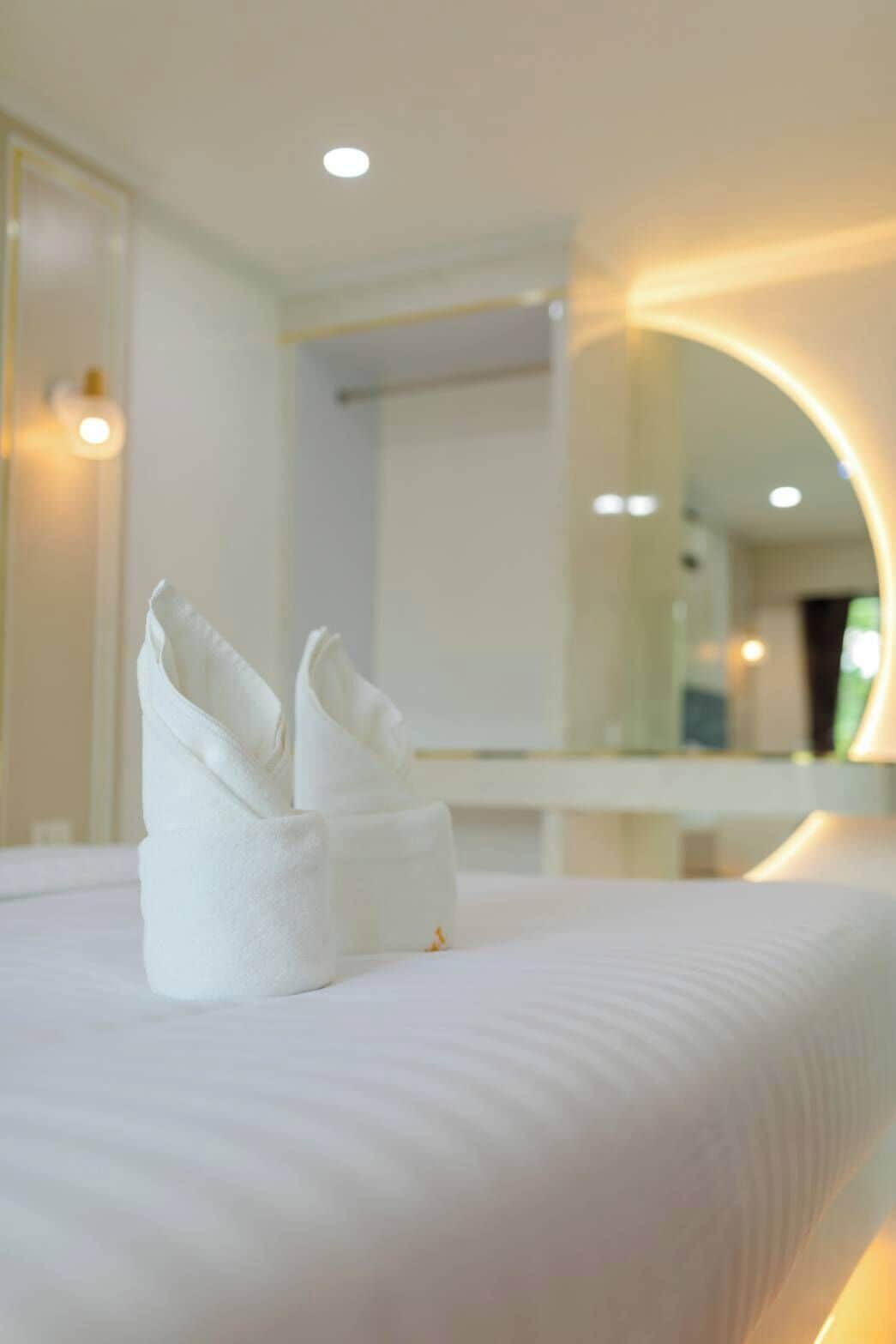 Un lit bien fait, avec une serviette pliée et une petite bouteille, probablement contenant de la lotion, dans une chambre d’hôtel moderne dotée d’un grand miroir et d’un éclairage encastré.