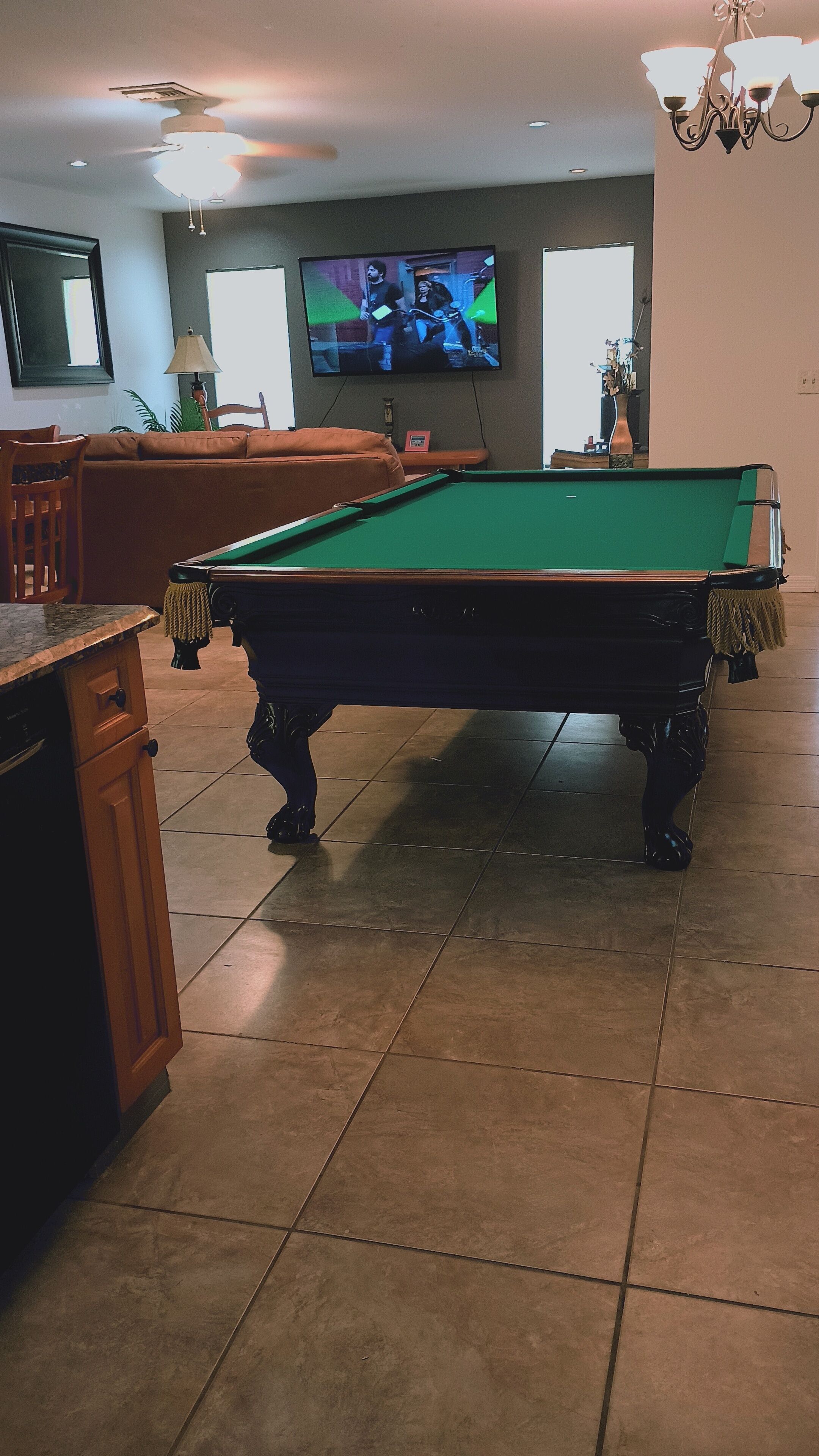 A pool table in a room with a television mounted on the wall, a ceiling fan, and a chandelier.