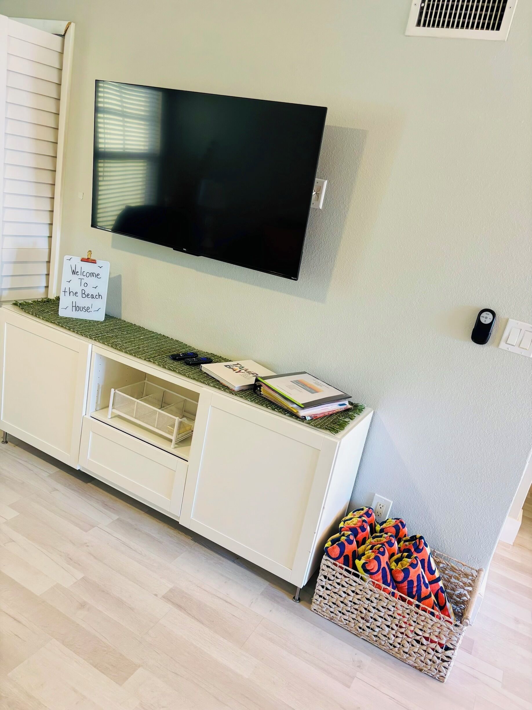 Una habitación con una televisión de pantalla plana montada en la pared, un armario blanco con un cartel que dice 'Welcome to the Beach House', una cesta de mimbre con objetos de colores y un enchufe en la pared.