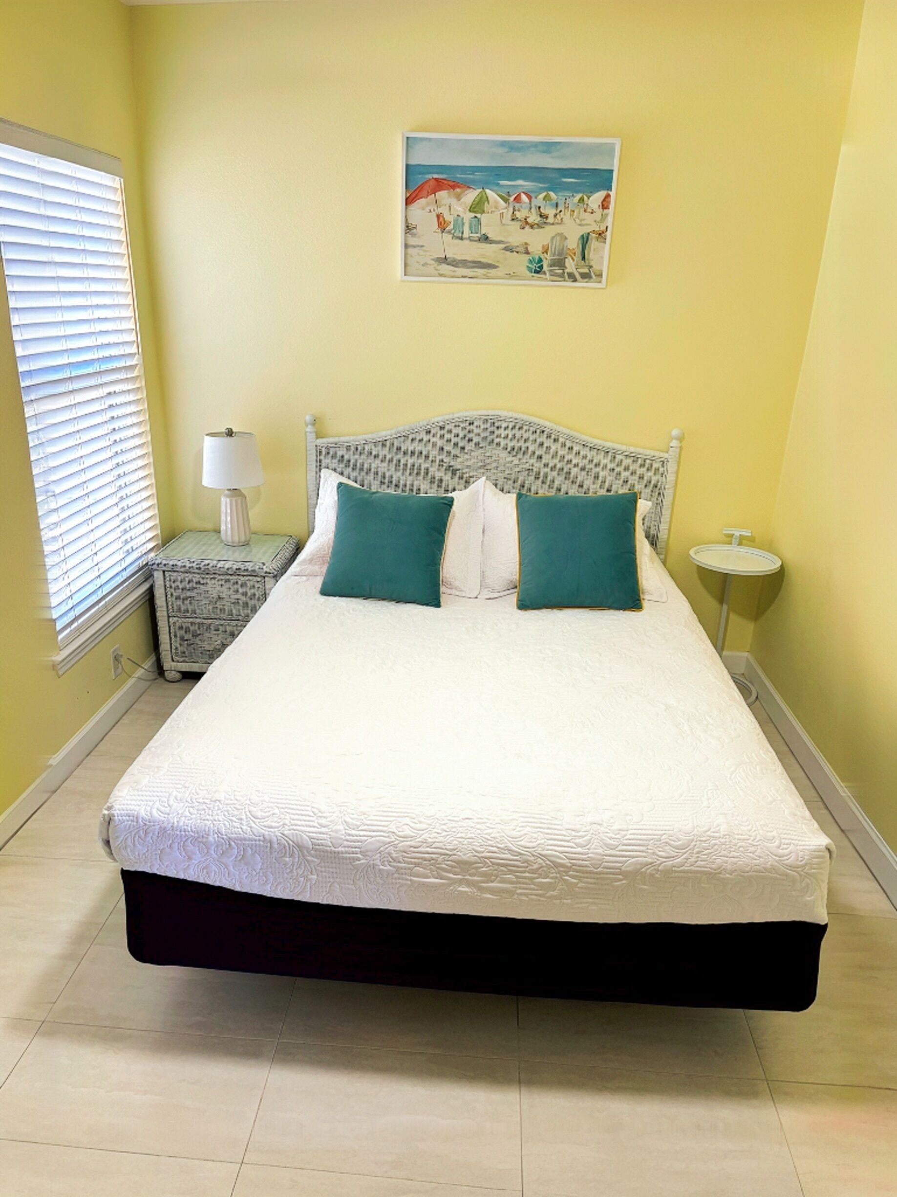 A bed with a white mattress and green pillows, a bedside table with a lamp, a painting of a beach scene, and a window with blinds.