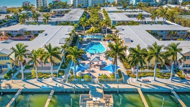 An aerial view of a resort with a central pool, palm trees, and a dock.