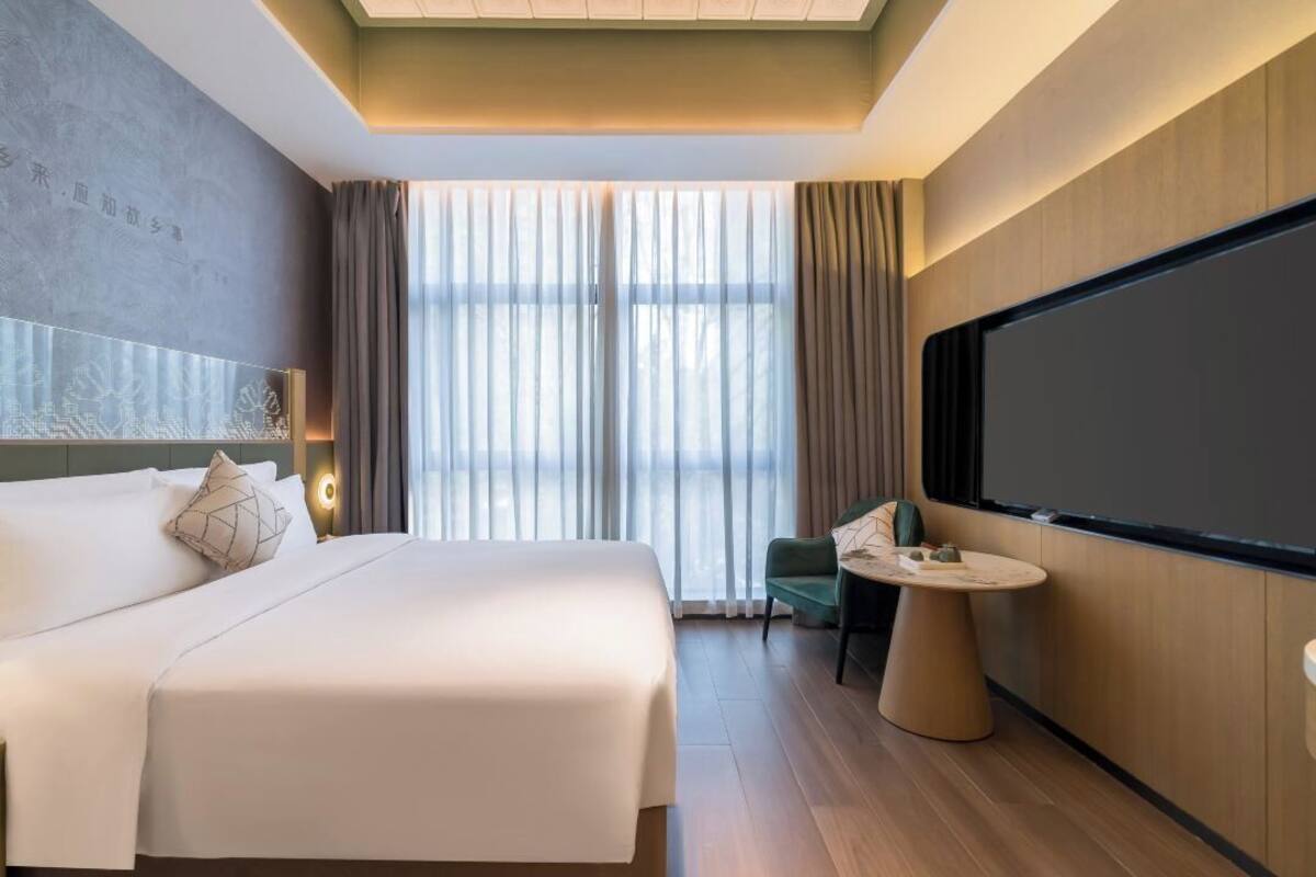 A modern hotel room with a large bed, a small round table, a television, and a view of the outdoors through sheer curtains.