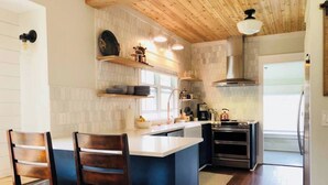A modern kitchen with a central island, wooden chairs, a built-in oven, and a stainless steel range hood. - The Perry Canary: Newly Renovated, Charming, Walk to Coffee and Restaurants (Spokane)