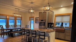 Una cocina moderna con encimeras de madera, taburetes de bar y una isla grande. Hay electrodomésticos de acero inoxidable, incluyendo un horno y una cafetera. La cocina tiene grandes ventanales que ofrecen vistas al exterior.