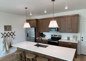 A modern kitchen with wooden cabinets, stainless steel appliances, a central island with stools, pendant lighting, and a painting on the wall. - Beautiful European Style Townhome - Near ISU (Pocatello)