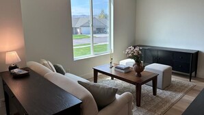 Un salon avec un canapé, une table basse, une lampe, un vase de fleurs et une vue sur une maison, visible à travers une grande fenêtre.