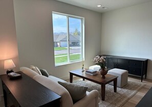 A living room with a sofa, a coffee table, a lamp, a vase with flowers, and a view of a house through a large window. - Beautiful European Style Townhome - Near ISU (Pocatello)