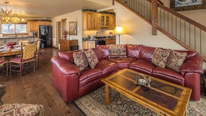 A spacious living room with a leather sectional sofa, a wooden coffee table, and a patterned rug. A staircase with wooden railings leads to an upper level.