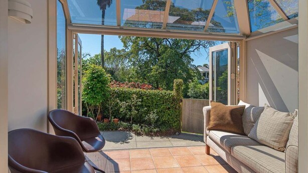 Eine sonnendurchflutete Terrasse mit Glastür zum Garten, einer gepolsterten Sitzecke und einem gefliesten Boden.