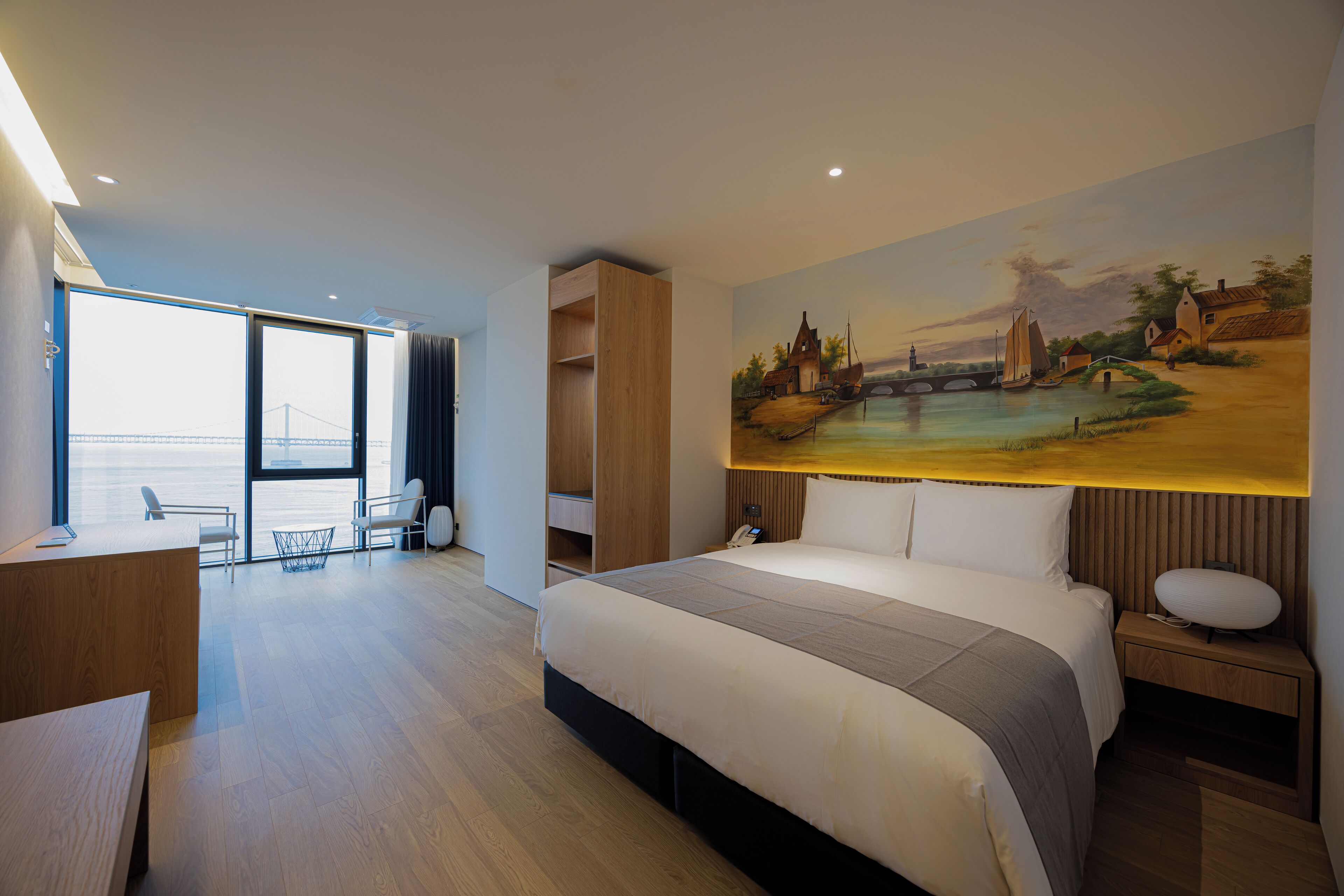 A modern hotel room with a large bed, a desk, and a painting of a landscape.