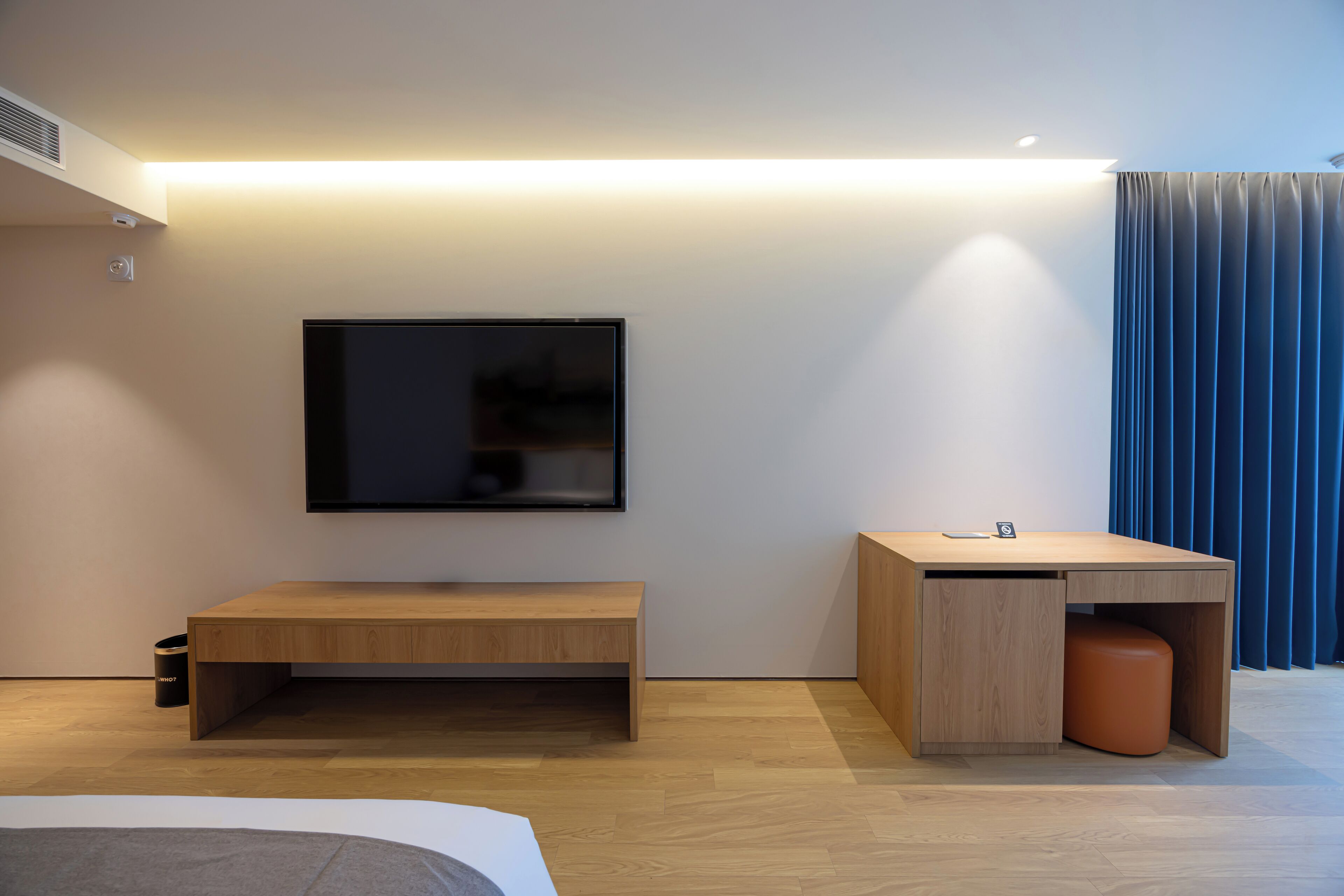 A modern hotel room with a wooden desk, a bench, a flat-screen TV mounted on the wall, and a window with blue curtains.