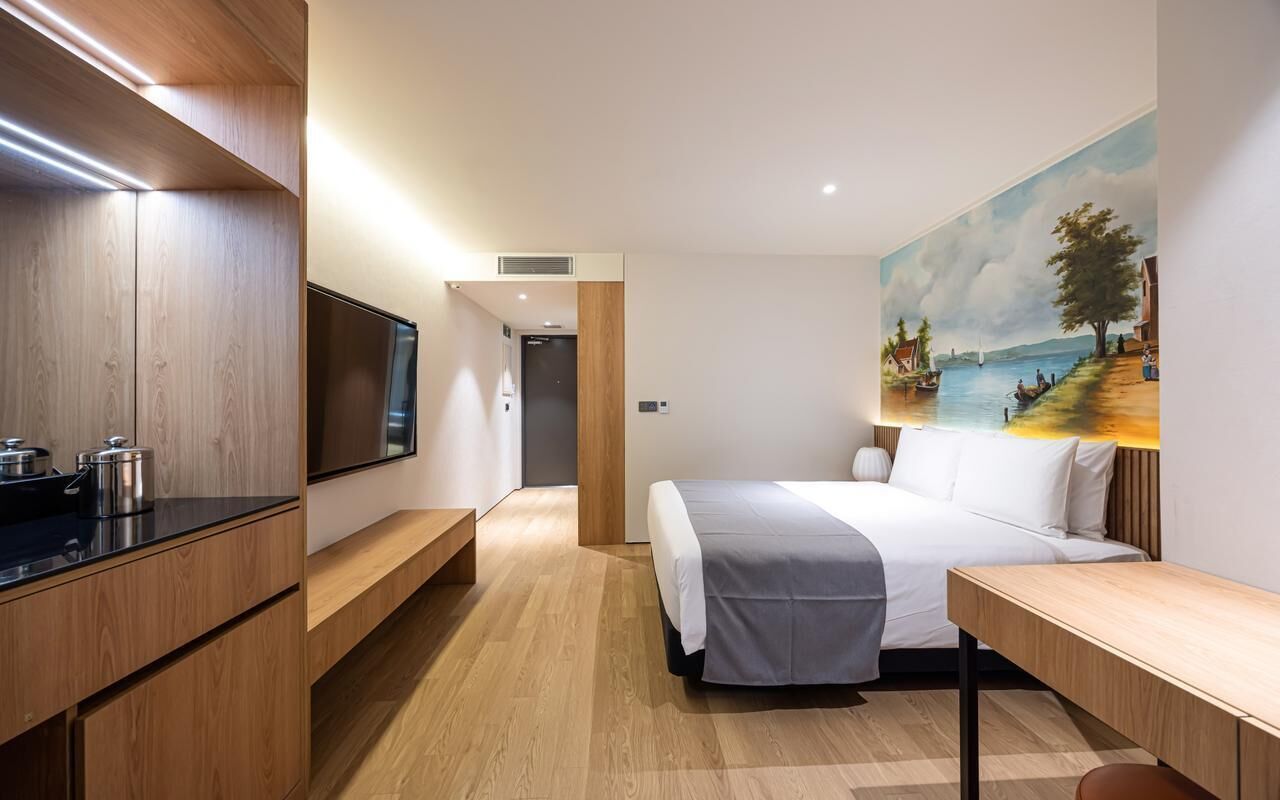 A modern hotel room with a bed, a TV, a painting, and a wooden desk.