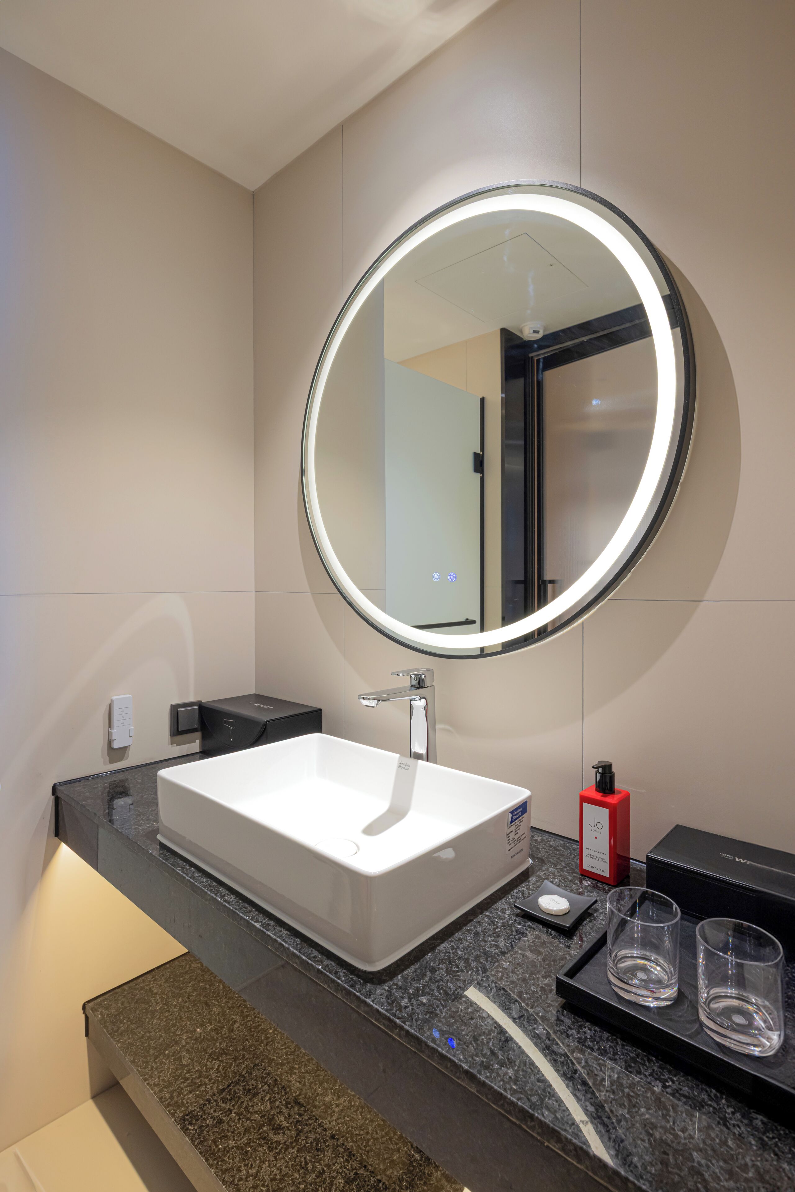 A modern bathroom with a white sink, a round mirror with a light, and a black countertop.