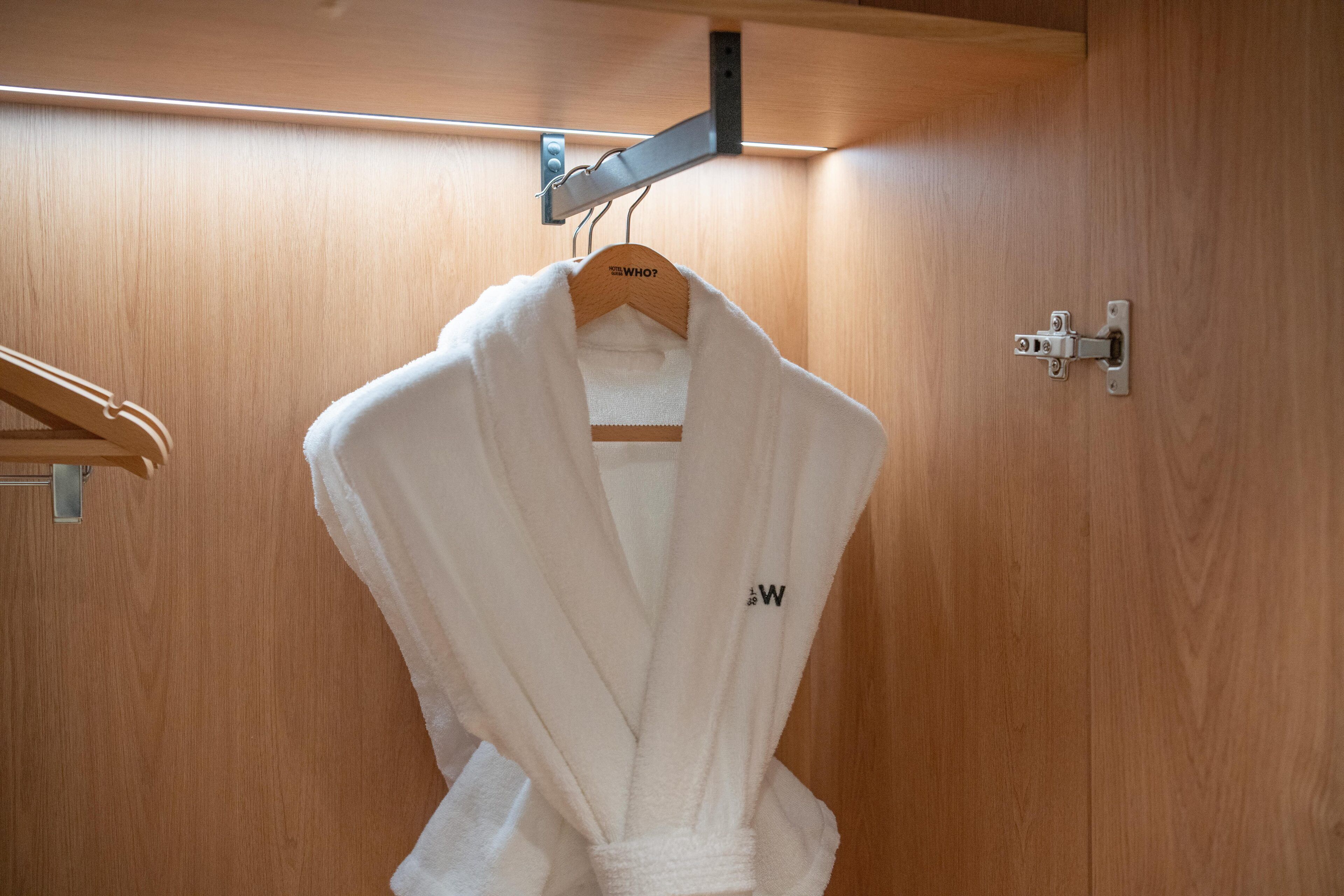 Une robe blanche est suspendue à un cintre en bois dans une pièce en bois, où l’on aperçoit un support métallique et une serrure de porte.