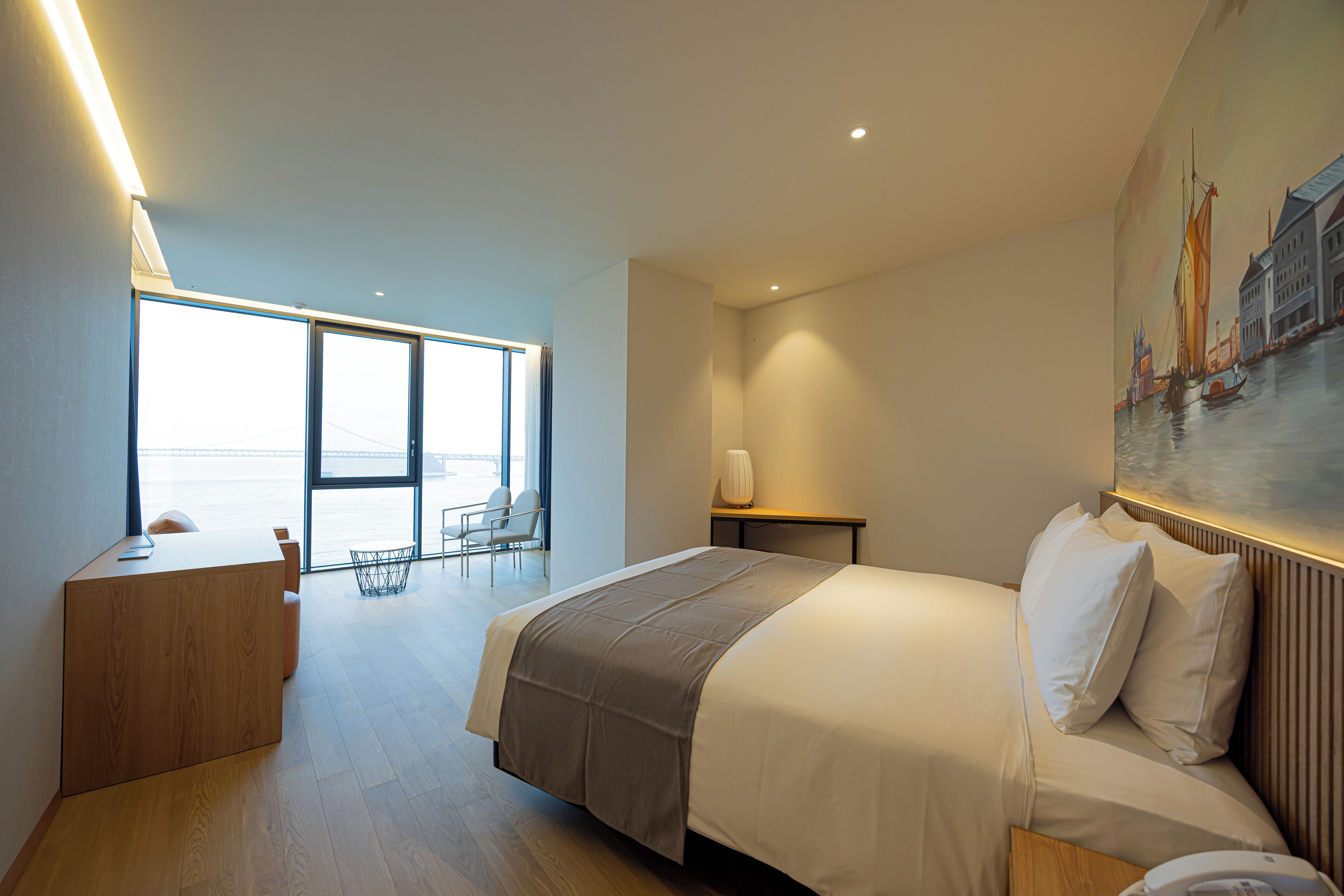 Habitació d'hotel moderna amb un llit gran, una taula de treball i vistes al mar a través de grans finestres.