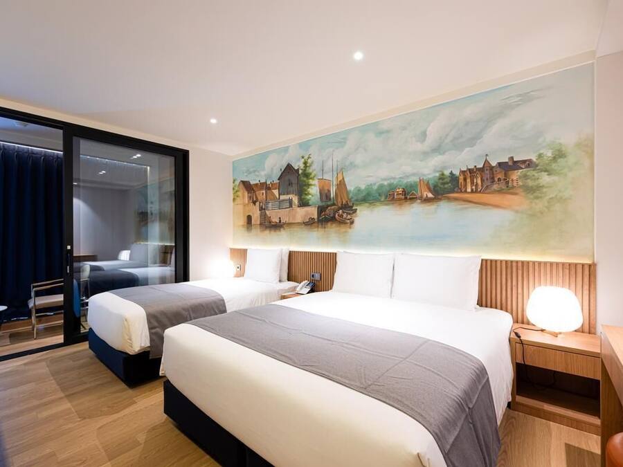 A hotel room with two beds, a painting of a riverside scene, a desk, and a chair.