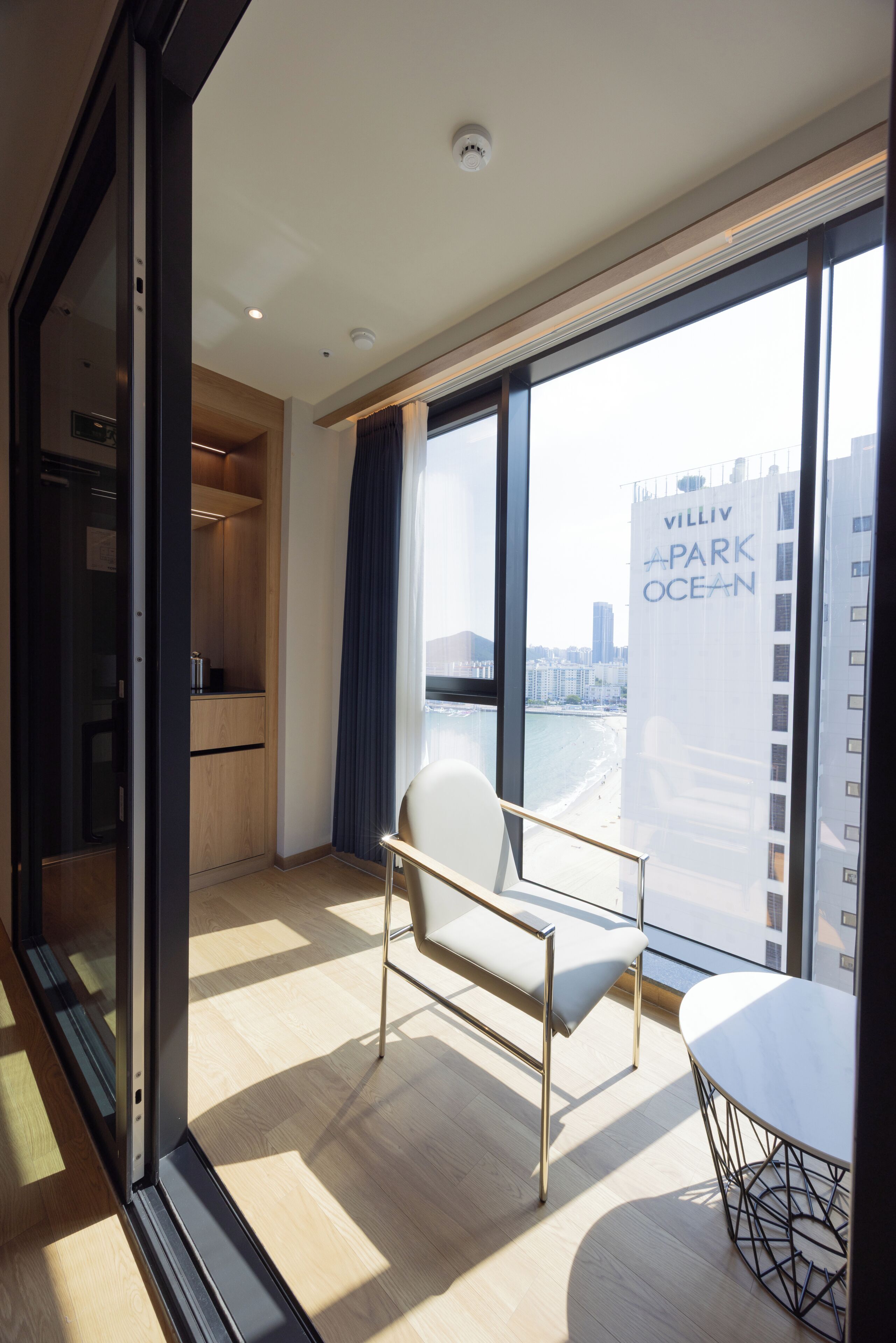 A modern hotel room with a large window, a seating area with a table, and a kitchenette.