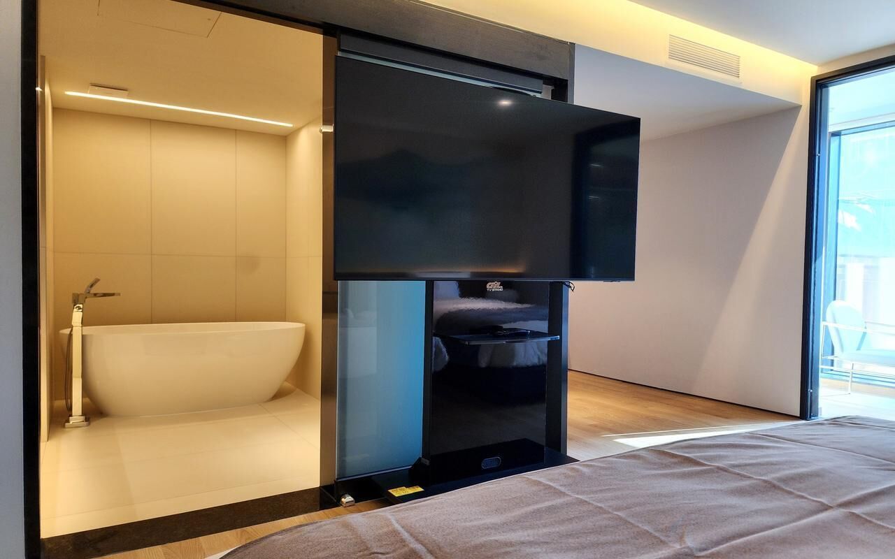A modern hotel room with a large bed, a built-in TV, a bathtub, and a sliding door leading to a balcony.