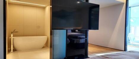 A modern hotel room with a large bed, a built-in TV, a bathtub, and a sliding door leading to a balcony.
