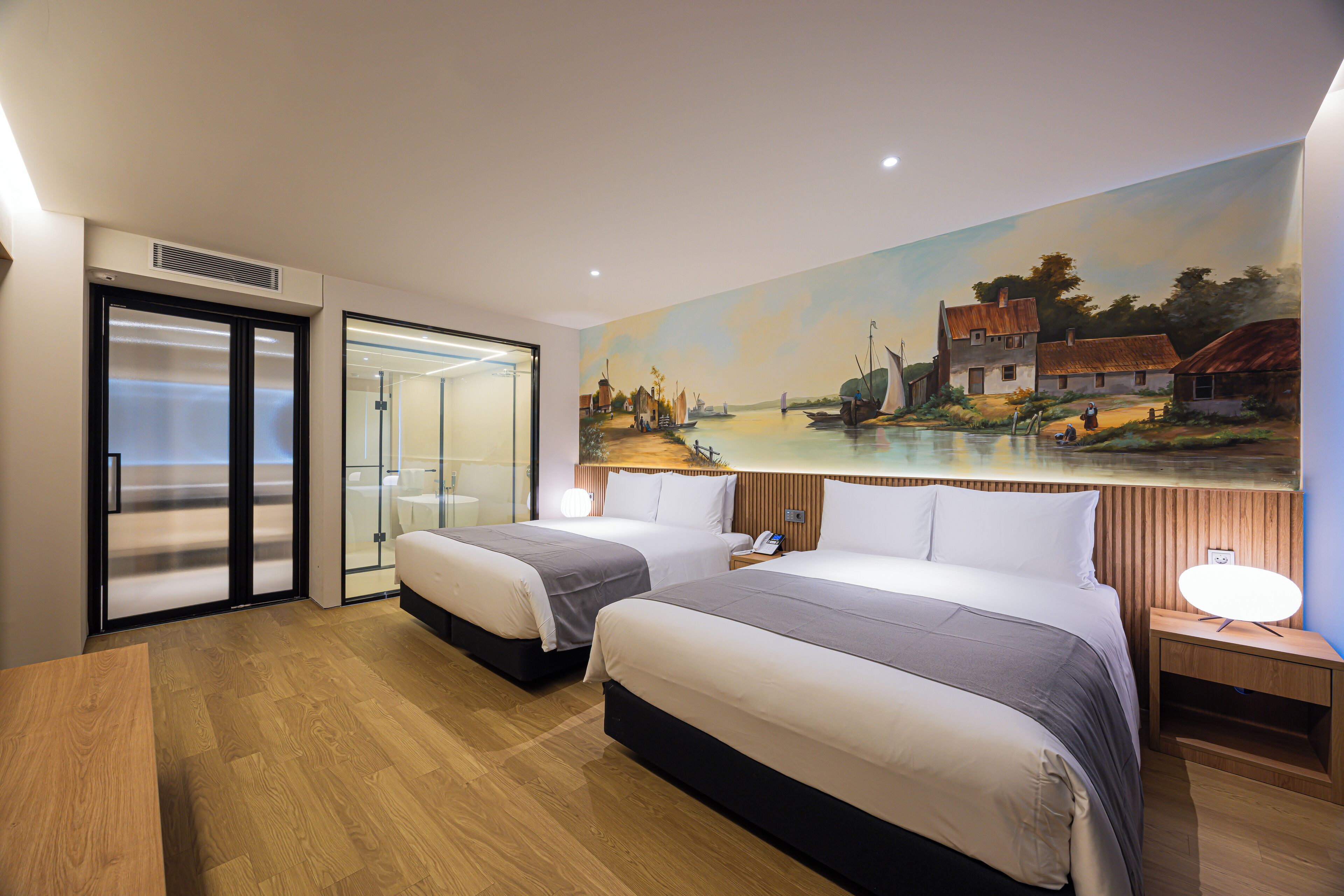 A hotel room with two beds, a painting of a village scene on the wall, a wooden floor, and a desk with a lamp.
