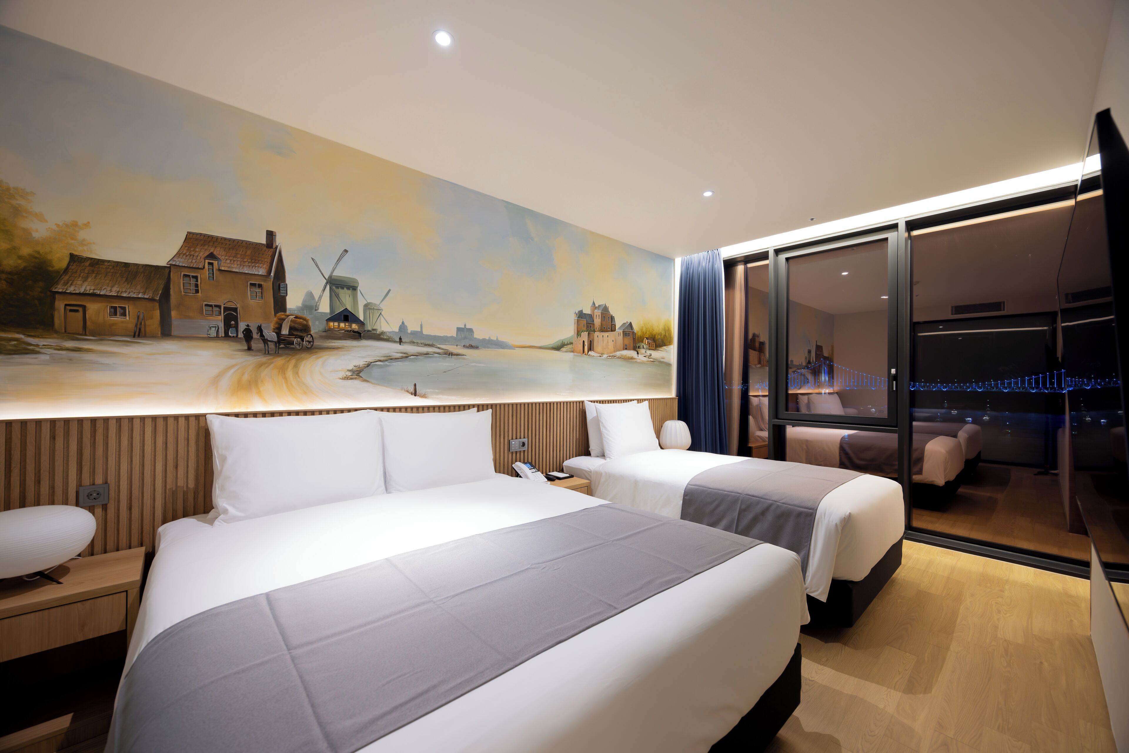 A hotel room with two beds, a large mural of a rural scene, and a view through a glass door.