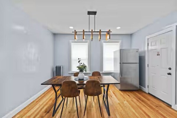 A modern kitchen with a wooden dining table, chairs, a refrigerator, and pendant lighting.