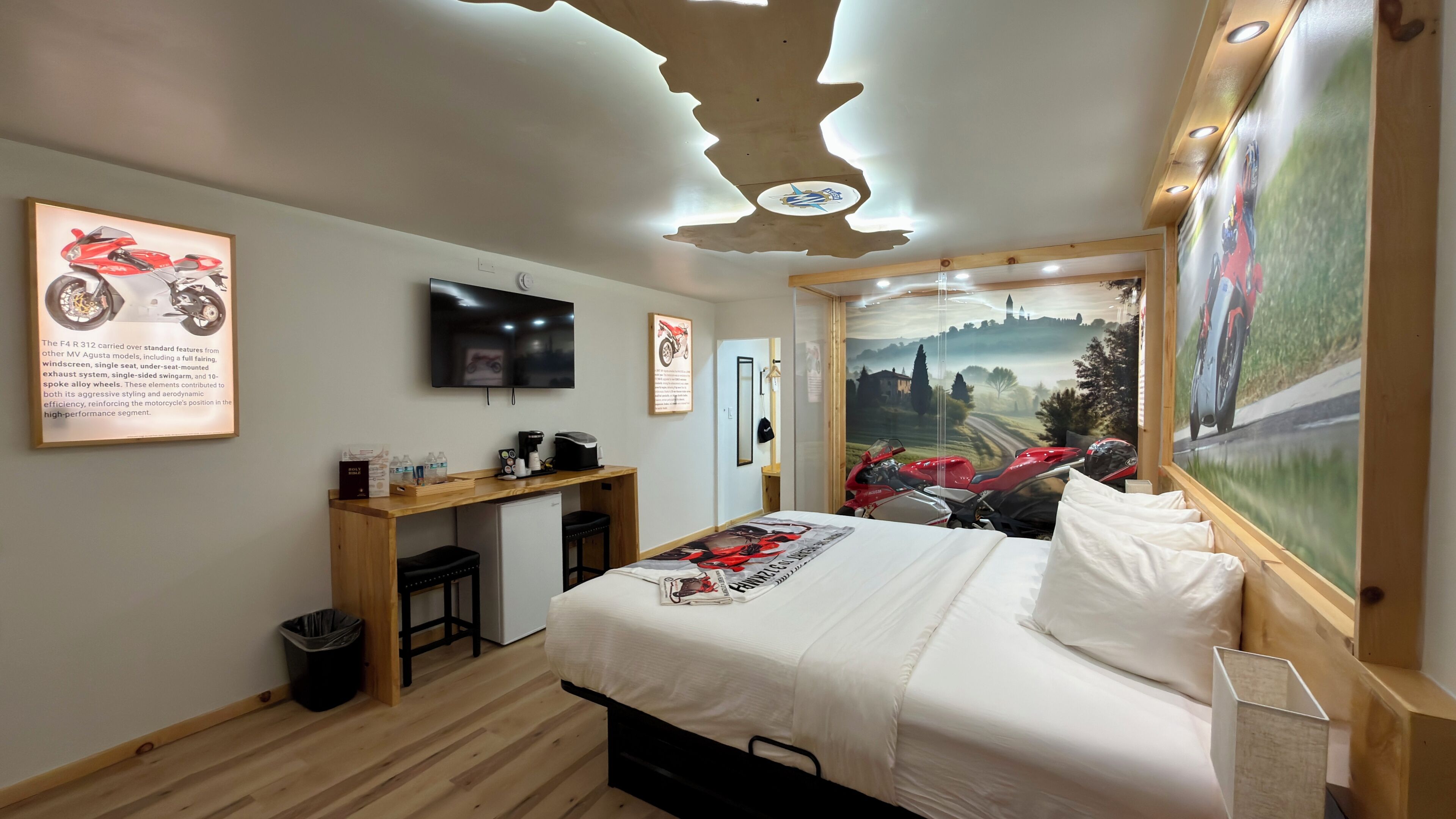 A hotel room with a bed, a desk with a computer, a TV, and a motorcycle-themed wall art.