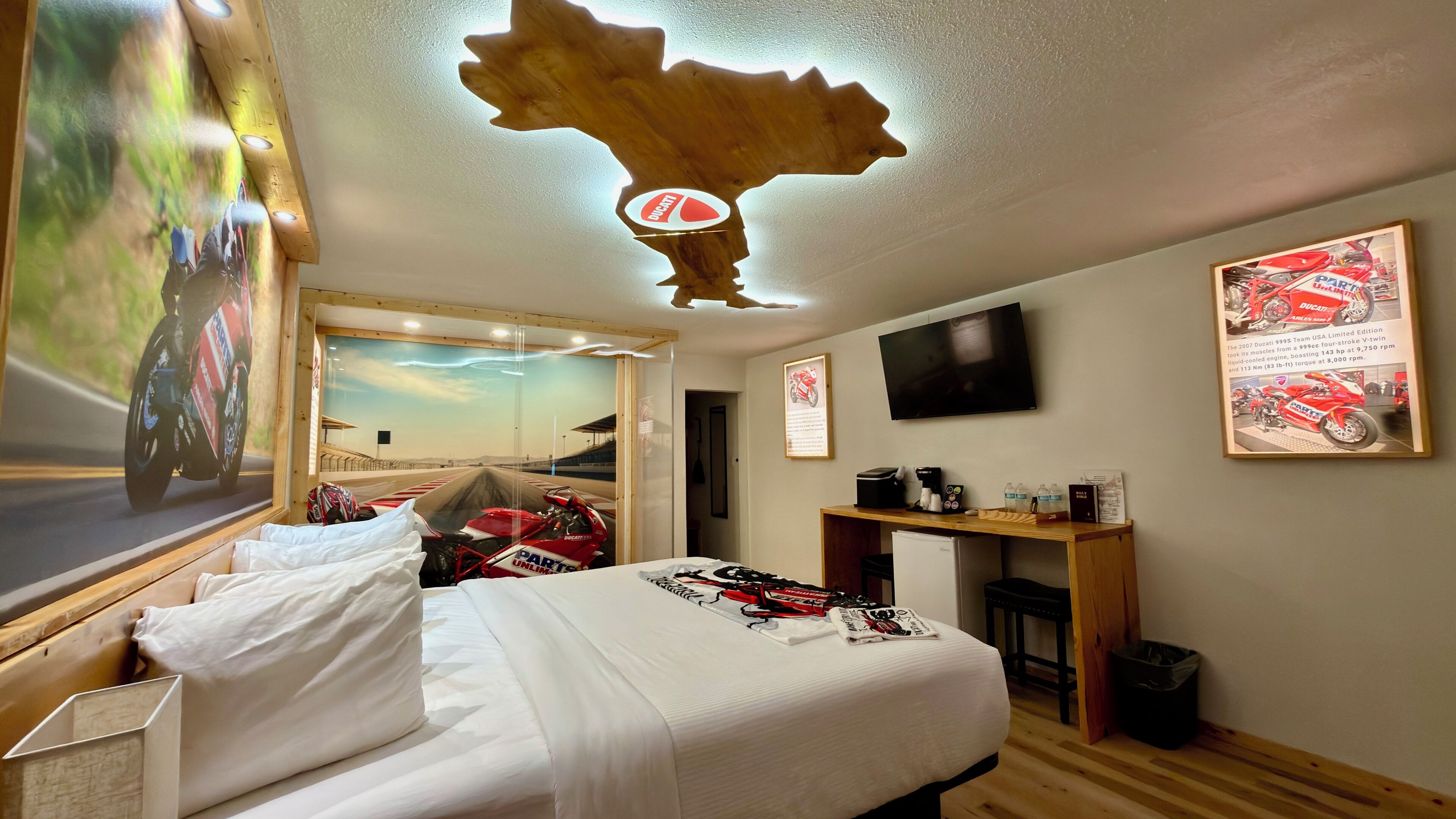 A hotel room with a bed, a mounted television, a wooden headboard, a wall-mounted mirror, and a framed motorcycle poster.