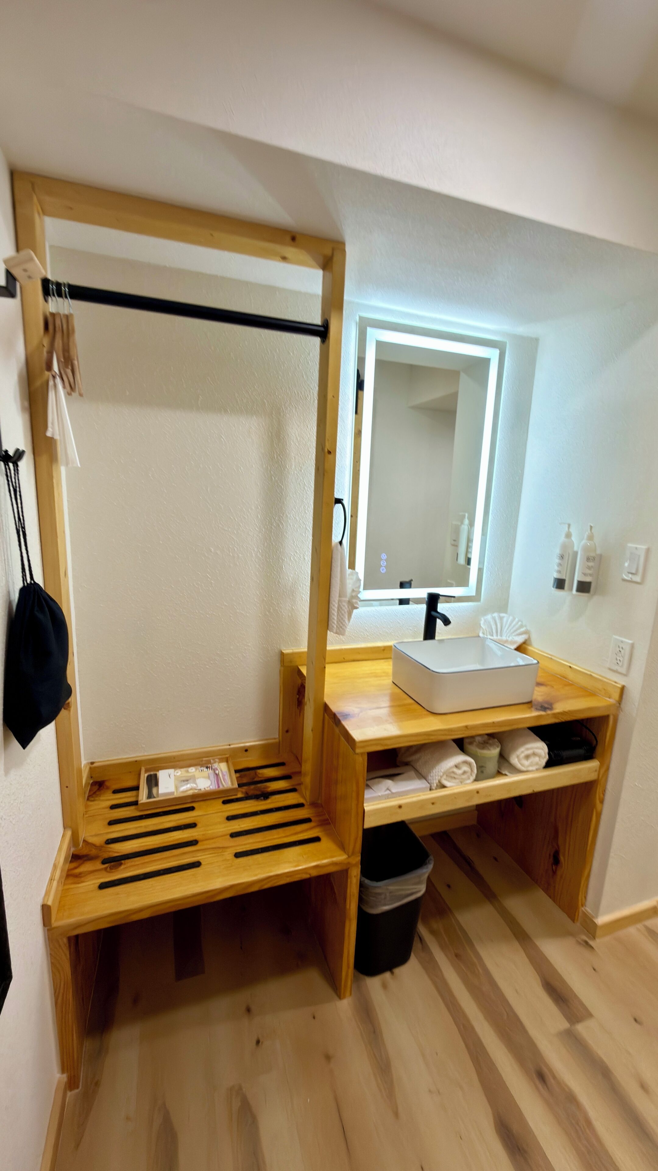 A compact bathroom with a wooden bench, a sink, a mirror, and a small trash bin.