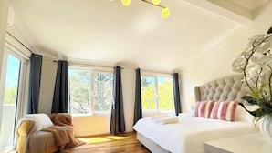 A bedroom with a large bed, a chair, and a desk. There are windows with curtains, a ceiling light, and a plant.