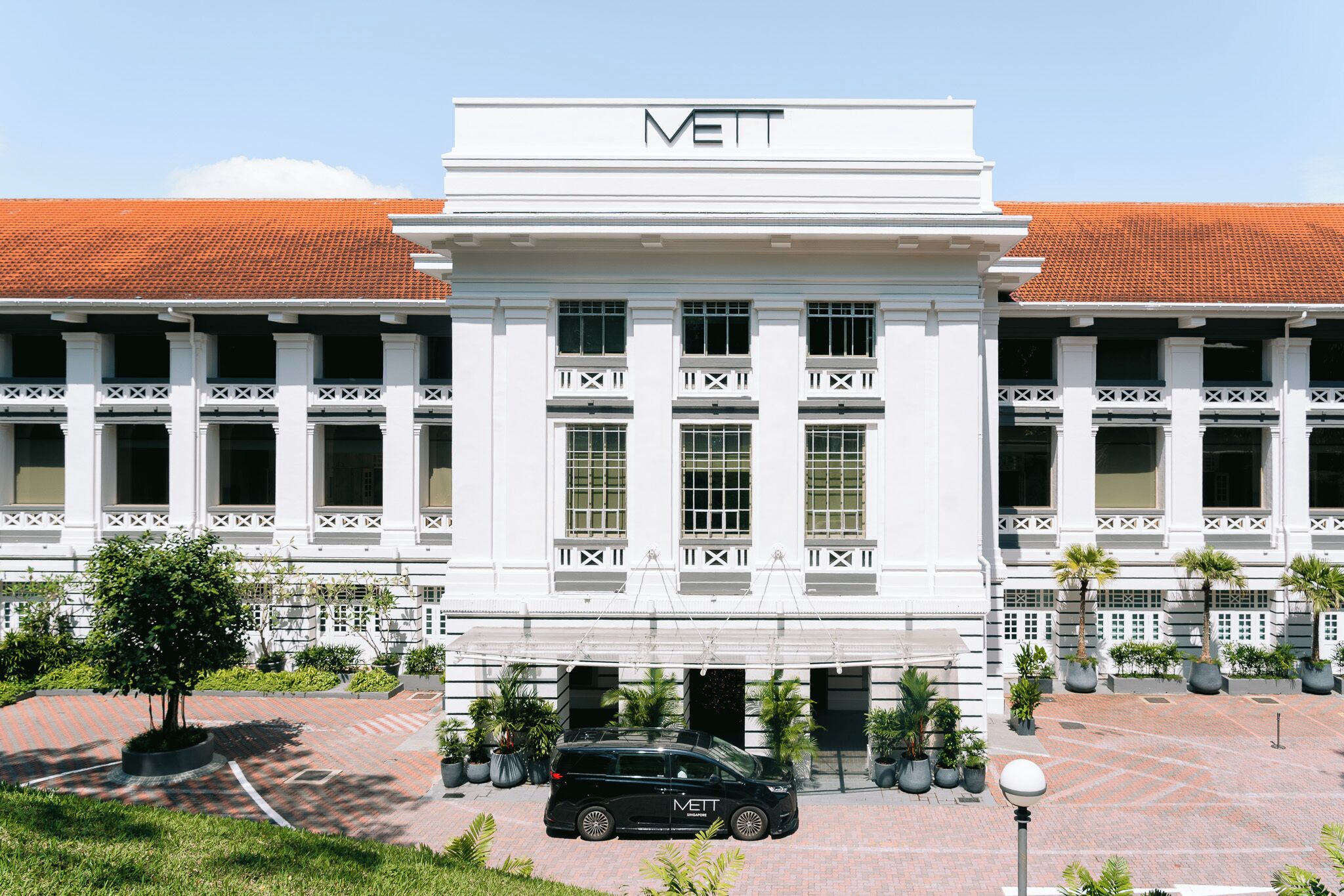 A white building with 'METT' on the facade, large windows, and a parking area with a black van.