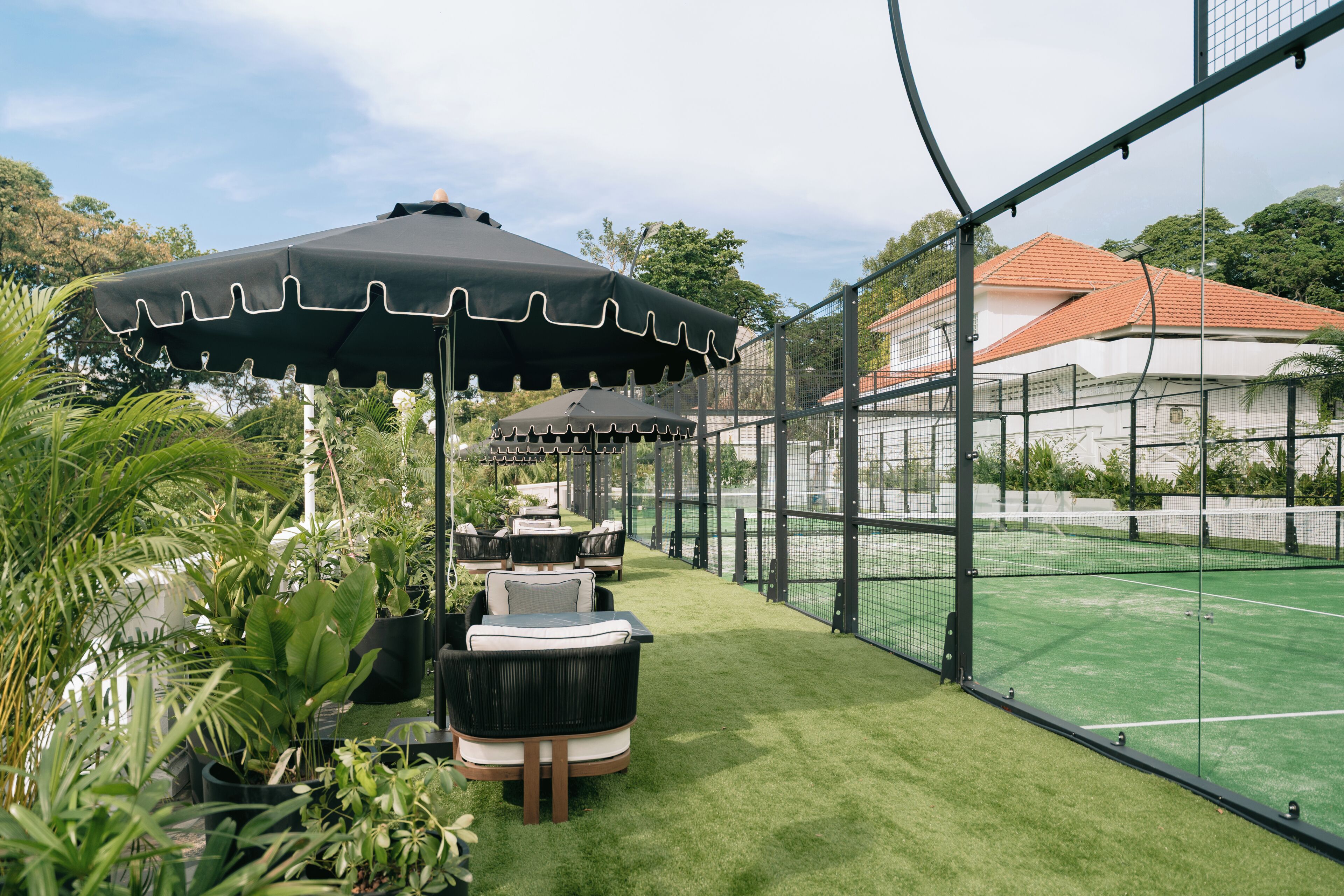 Een tennisbaan met een net, omringd door een hek, en een zithoek met stoelen en parasols.