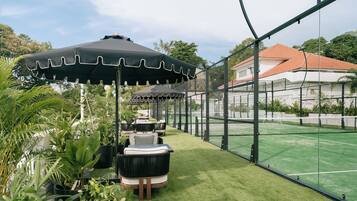 Een tennisbaan met een net, omringd door een hek, en een zithoek met stoelen en parasols.