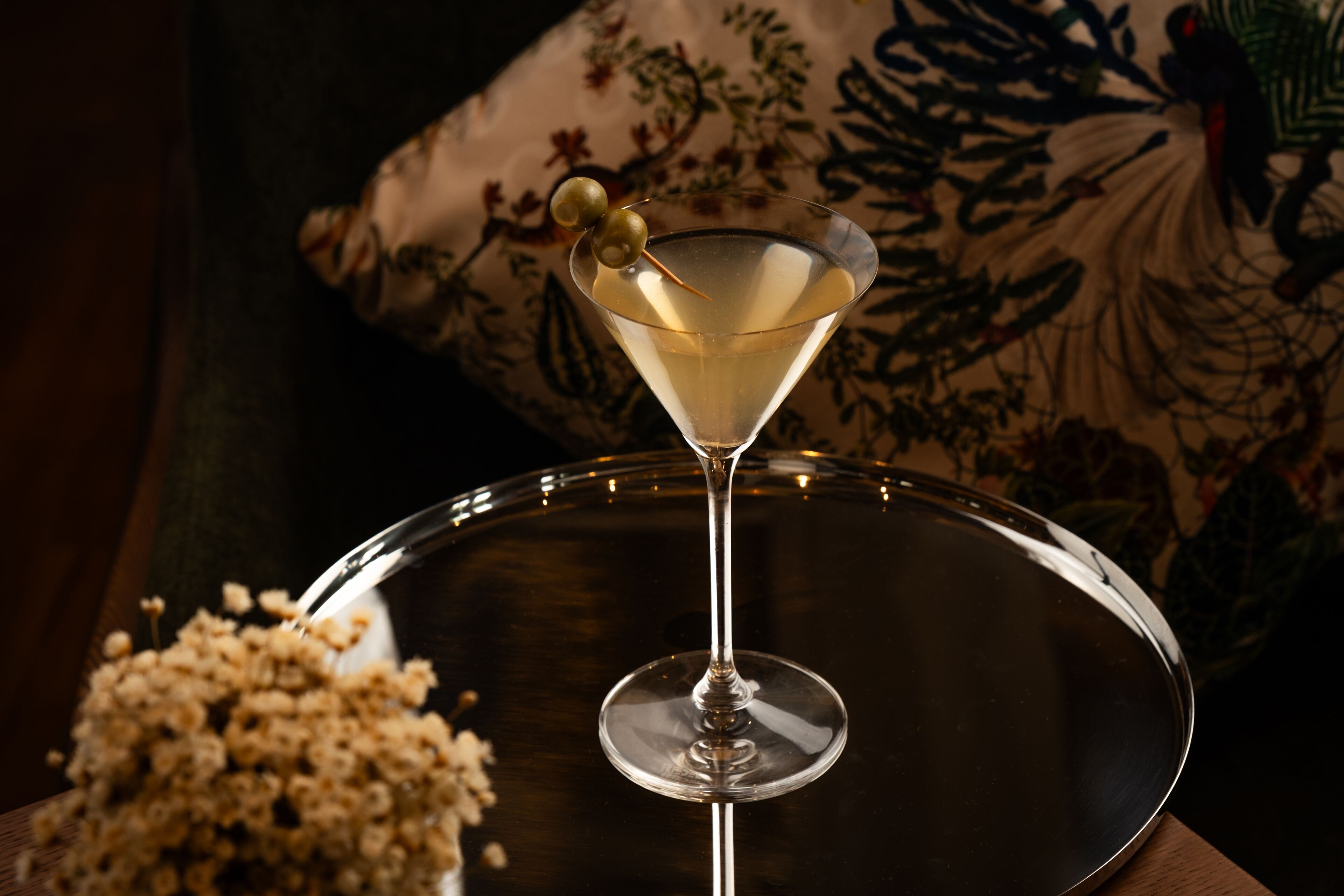 Een martini glas met garnering op een reflecterend dienblad, voor een kussen met bloemenmotief.