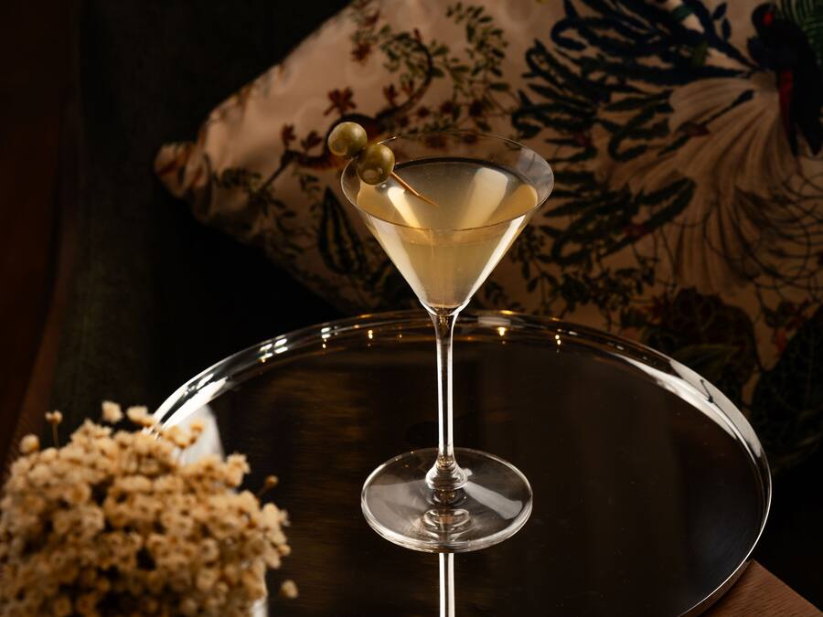 Een martini glas met garnering op een reflecterend dienblad, voor een kussen met bloemenmotief.