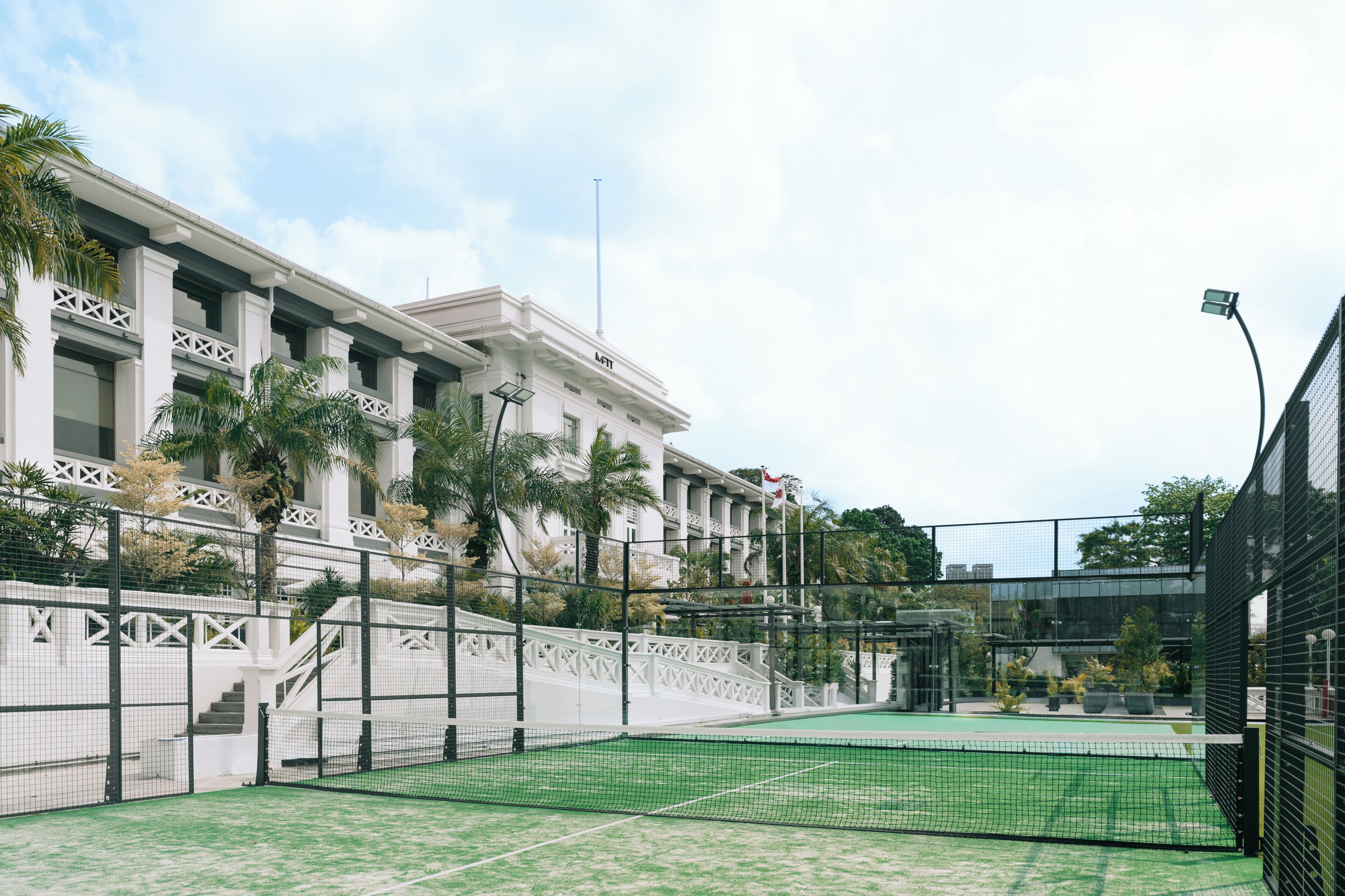 A tennis court with a net, surrounded by a white fence and palm trees.