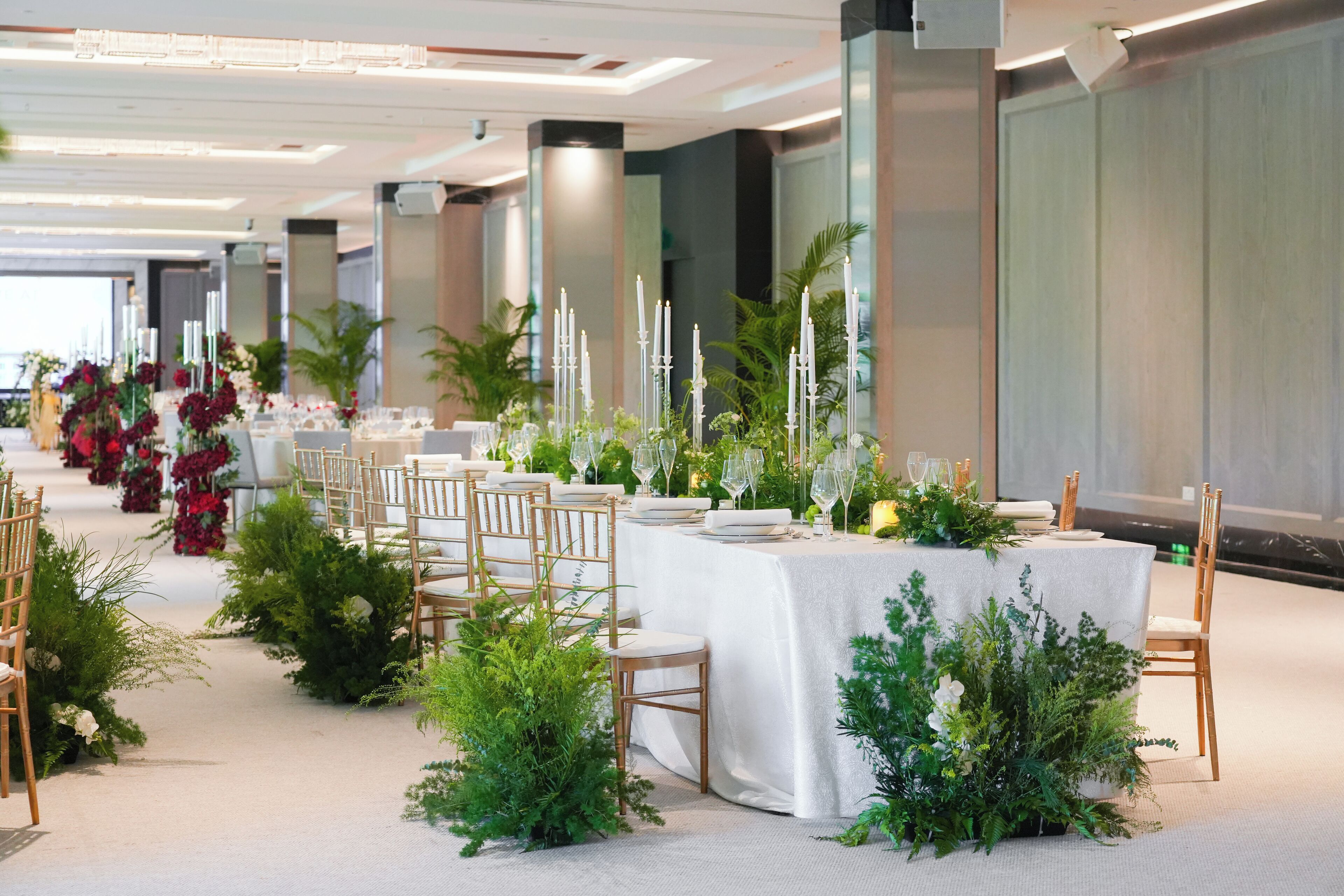 Un salón de banquetes con largas mesas dispuestas para un evento formal, rodeado de follaje y velas.