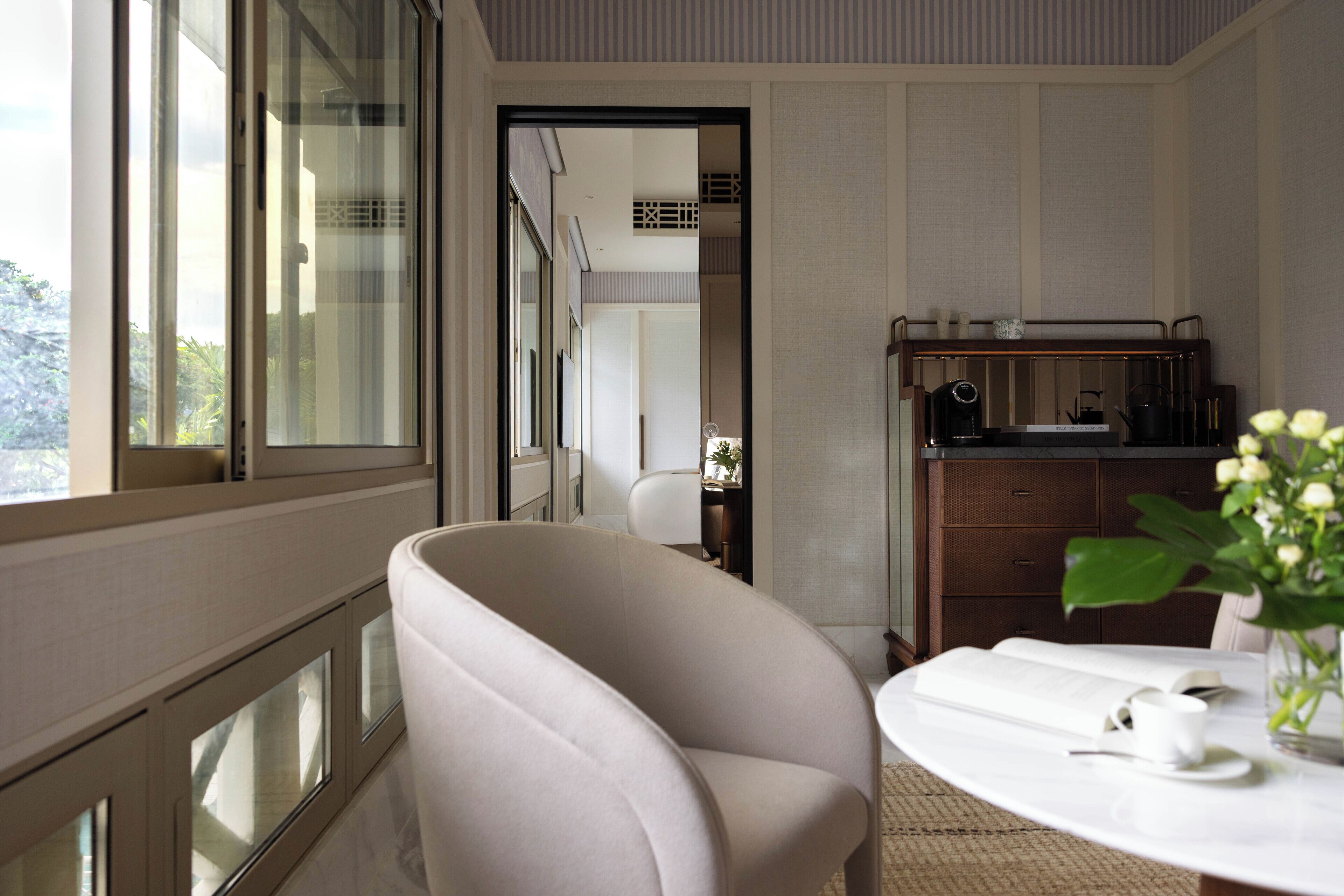 Una habitación moderna con un ventanal grande, un sillón blanco, una mesa redonda con un libro y una taza, y un mueble pequeño con una radio.