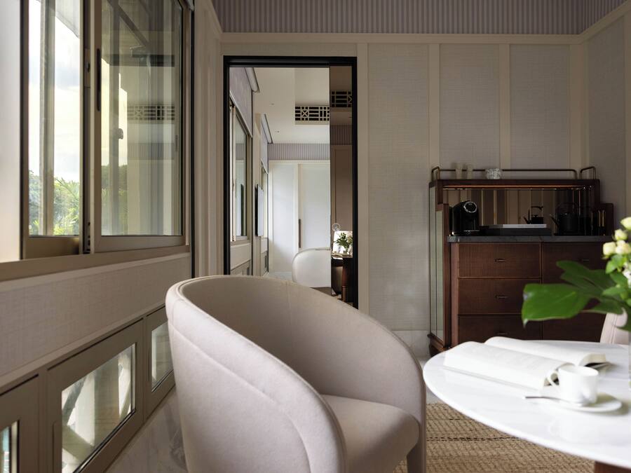 Una habitación moderna con un ventanal grande, un sillón blanco, una mesa redonda con un libro y una taza, y un mueble pequeño con una radio.